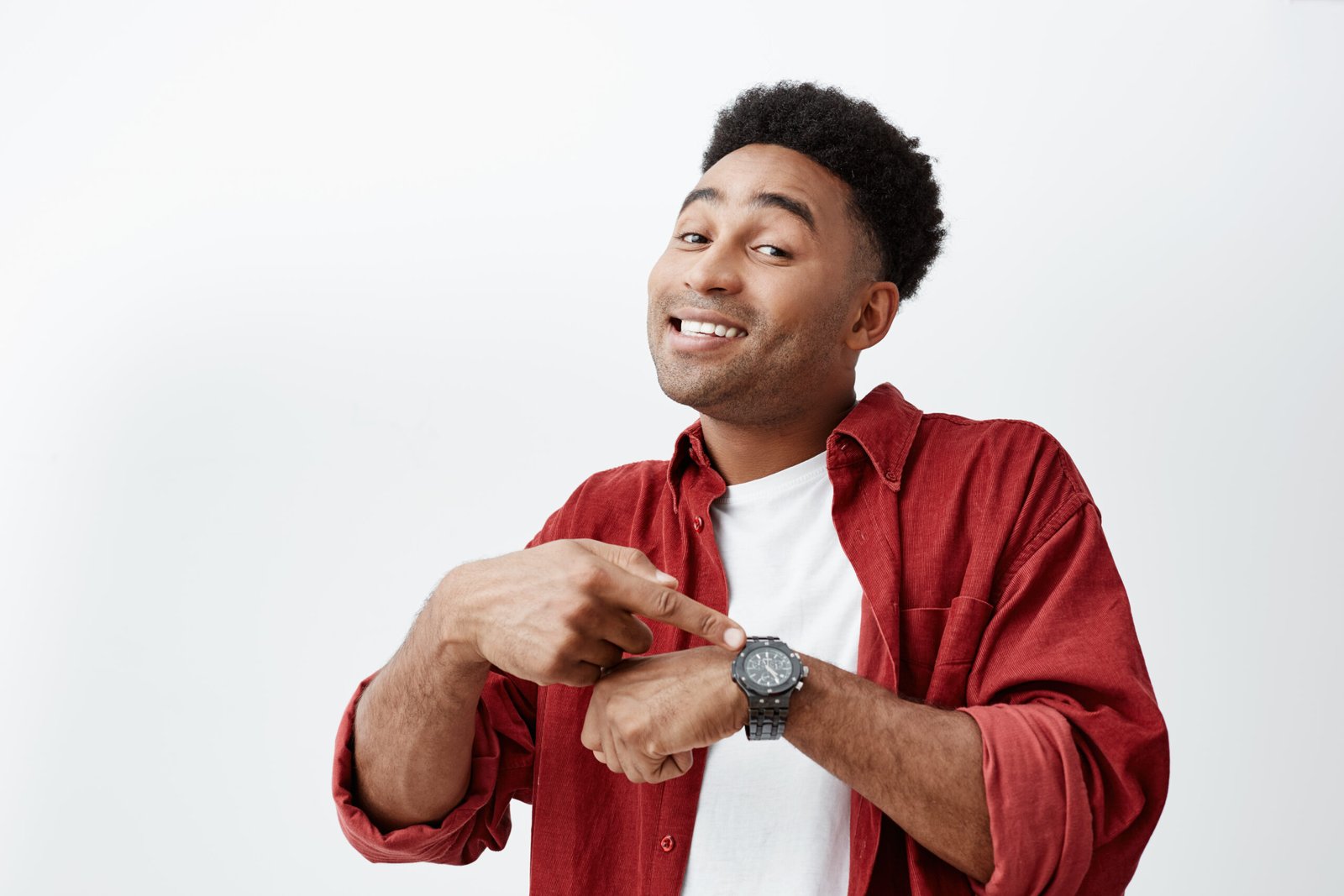 What time is it. Portrait of young attractive dark-skinned man with dark afro hairstyle in white t-shirt and red shirt pointing at hand watch with happy face expression, showing it time to eat
