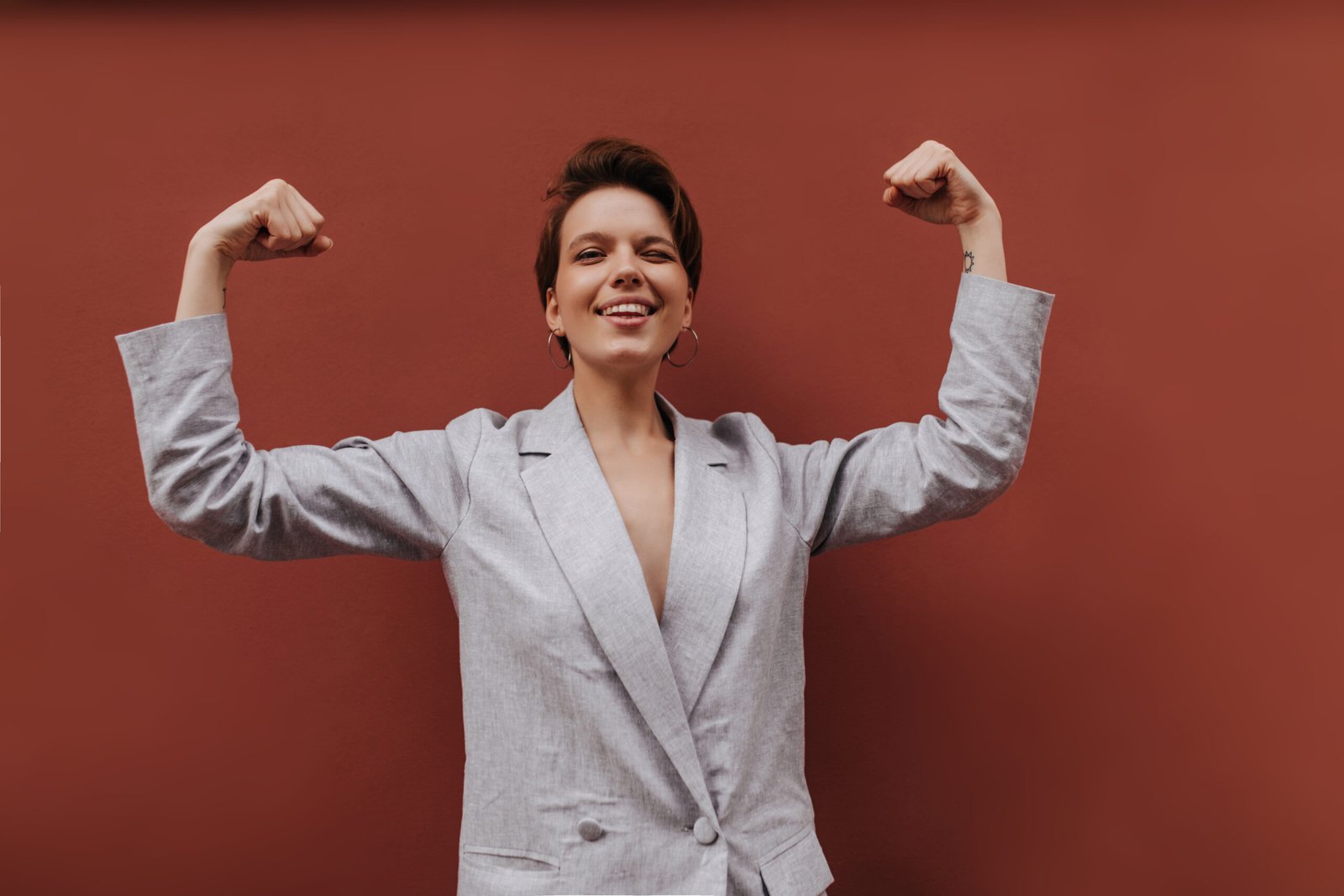 Pretty lady in suit showing her biceps on brown background. Short-haired woman in grey stylish jacket demonstrates power on isolated